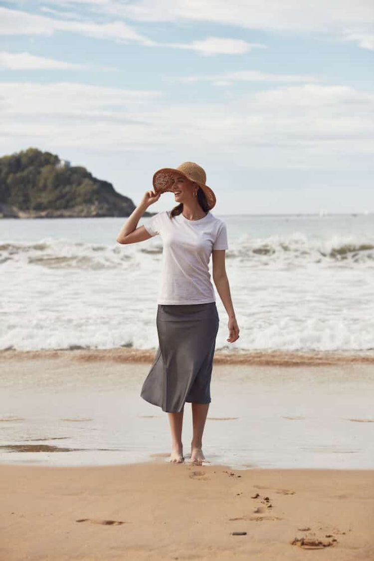 Woman standing on a beach wearing a white shirt, gray skirt, and straw hat.